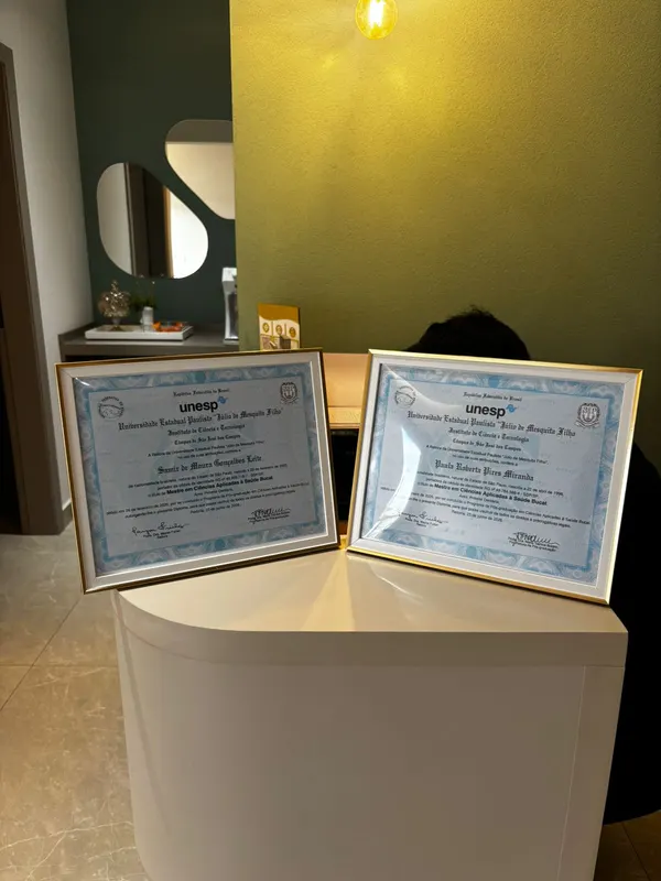 Os dois diplomas de mestrado da UNESP emoldurados na estante da clínica